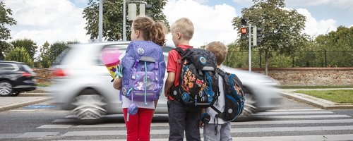 3 Kinder warten am Gehsteig vor einem Zebrastreifen - ein Auto fährt gerade vorbei.