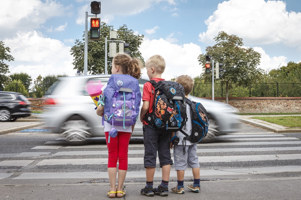 3 Kinder warten am Gehsteig vor einem Zebrastreifen - ein Auto fährt gerade vorbei.
