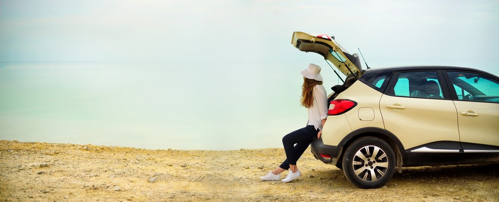 helles Auto am Meer. Frau lehnt am geöffneten Kofferraum und blickt aufs Meer.