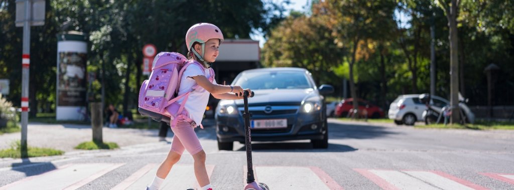 Schulkind ueberquert mit Scooter und Schultasche am Ruecken einen Zebrastreifen